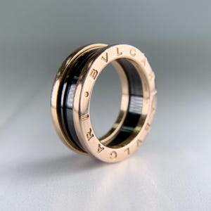 May include: A gold-tone ring with a black band. The ring features the brand name engraved around the outer edge. The design is a combination of gold and black, creating a modern and elegant look. The ring is a piece of jewelry.