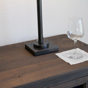 May include: A dark brown wooden table top with a black lamp and a clear wine glass on a white linen napkin.