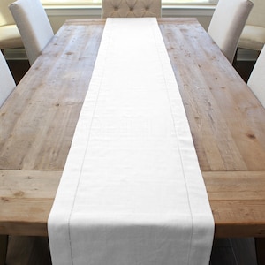 May include: A white linen table runner with a subtle texture and a stitched edge. The runner is laid on a wooden table with chairs on either side.