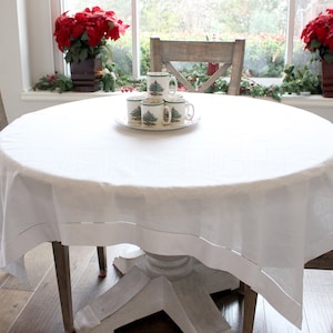 May include: A white round tablecloth with a delicate lace trim, set on a round wooden table with four chairs. The table is decorated with a plate of five white mugs with a green Christmas tree design.