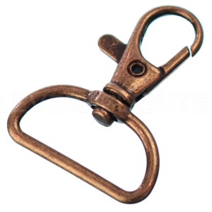 May include: A bronze-colored metal swivel clasp with a D-ring. The clasp is open and ready to be attached to a strap or chain.