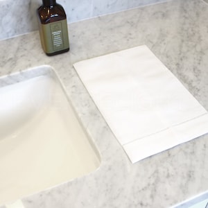 May include: A white hand towel with a subtle embroidered border sits on a marble countertop next to a white porcelain sink.