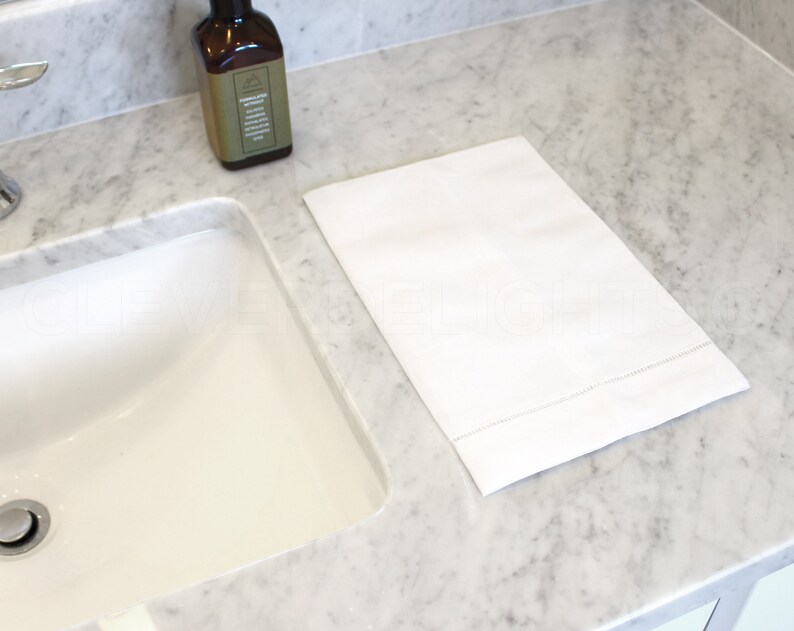 May include: A white hand towel with a subtle embroidered border sits on a marble countertop next to a white porcelain sink.