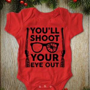 May include: Red baby bodysuit with the text "YOU'LL SHOOT YOUR EYE OUT" in black. The design includes a pair of glasses with a shattered lens and two rifles. The bodysuit is short-sleeved and has snap closures.
