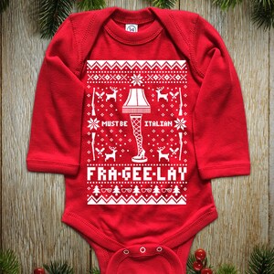 May include: Red baby bodysuit with long sleeves. The white graphic design includes a leg lamp, deer, rifles, and the text "MUST BE ITALIAN FRA-GEE-LAY". The bodysuit has a round neckline and snap closures at the bottom.
