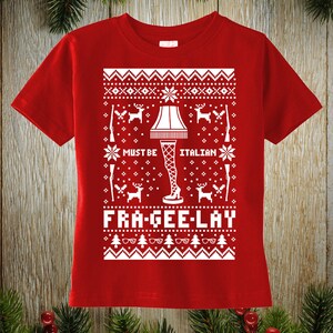 May include: Red t-shirt with a white graphic design. The design features a leg lamp, snowflakes, deer, and the text "MUST BE ITALIAN FRA-GEE-LAY". The shirt has a crew neck and short sleeves.