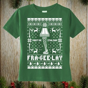 May include: Green t-shirt with a white graphic design. The design features a leg lamp, snowflakes, deer, and the text "MUST BE ITALIAN" and "FRA-GEE-LAY". The shirt has short sleeves and a crew neck.