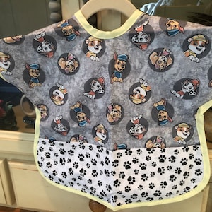 Reversible baby bib featuring a gray background with cartoon dog characters in gray circles. The bib has a white bottom section with black paw prints and is trimmed with yellow.