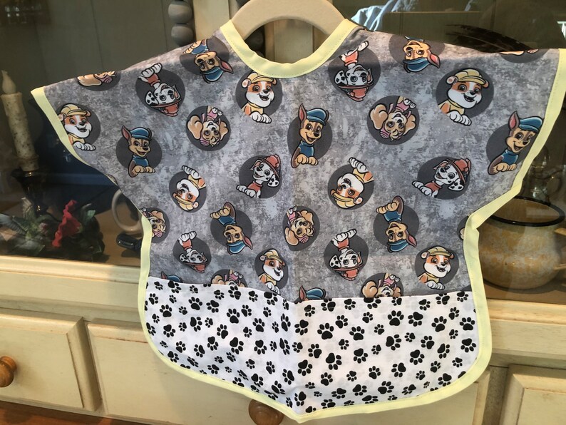 Reversible baby bib featuring a gray background with cartoon dog characters in gray circles. The bib has a white bottom section with black paw prints and is trimmed with yellow.