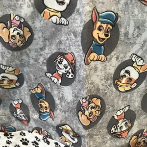 Gray fabric featuring cartoon images of the Paw Patrol characters in gray circles. The fabric has a textured background and a border of white fabric with black paw prints. The Paw Patrol characters are in various poses.