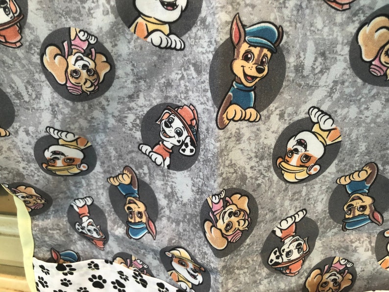 Gray fabric featuring cartoon images of the Paw Patrol characters in gray circles. The fabric has a textured background and a border of white fabric with black paw prints. The Paw Patrol characters are in various poses.