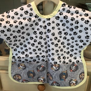 May include: A baby bib with a white background covered in black paw prints. The bottom section features a gray fabric with cartoon dog characters. The bib has a yellow trim and a pocket.