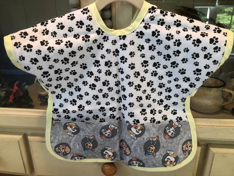 May include: A baby bib with a white background covered in black paw prints. The bottom section features a gray fabric with cartoon dog characters. The bib has a yellow trim and a pocket.