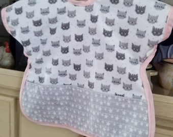 May include: A baby bib with a white background covered in gray cat faces. The bottom portion features a gray star pattern. The bib is trimmed in pink and hangs from a wooden hanger.