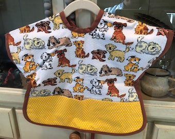 May include: A baby bib with a white background covered in various cartoon dog illustrations. The bib has a brown trim and a yellow pocket with white polka dots. It is hanging on a white hanger.