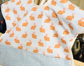May include: A baby bib with a light blue background covered in orange bunny illustrations. The bottom section features a light blue fabric with small white flowers. The bib has a white trim and ties at the back.