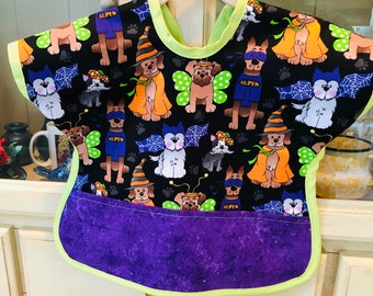 May include: A Halloween-themed baby bib with a black background featuring cartoon dogs in costumes, including witch hats, capes, and superhero outfits. The bib has a purple pocket, a lime green trim, and the word "SUPER" on some of the dog's outfits.