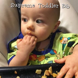 May include: A baby wearing a green and blue patterned bib is eating food. The bib has a blue trim and the text "Supreme Toddler Bib" is at the top. The baby is in a high chair and has food on their face and hands.