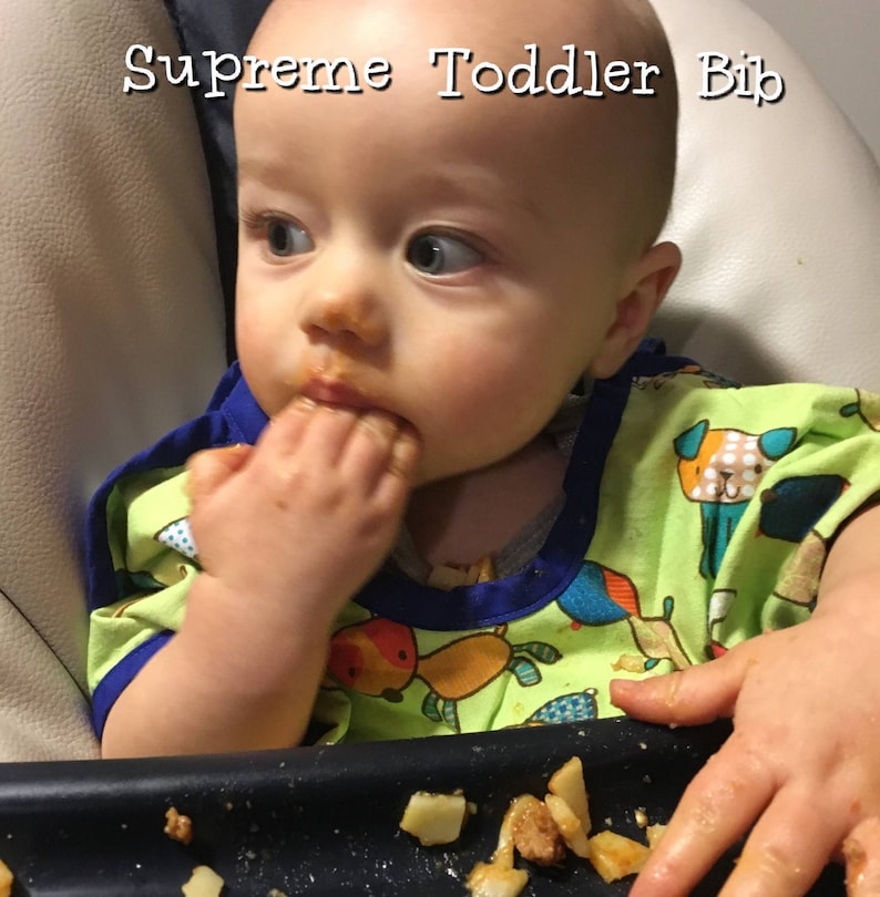 May include: A baby wearing a green and blue patterned bib is eating food. The bib has a blue trim and the text "Supreme Toddler Bib" is at the top. The baby is in a high chair and has food on their face and hands.