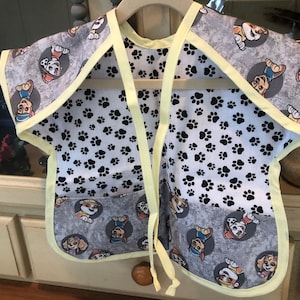 May include: A gray and white bib with a yellow trim and ties. The bib features a pattern of cartoon dog characters in gray circles and black paw prints. The bib is designed to protect clothing during mealtime.