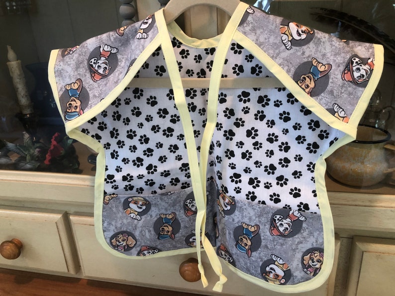 May include: A gray and white bib with a yellow trim and ties. The bib features a pattern of cartoon dog characters in gray circles and black paw prints. The bib is designed to protect clothing during mealtime.