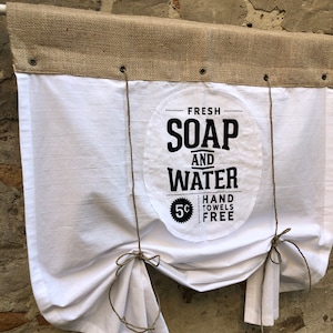 Könnte beinhalten: Ein weißer Stoff-Vorhang mit einer Jute-Oberseite und einem schwarz-weißen Schild mit der Aufschrift "Fresh Soap and Water 5¢ Hand Towels Free".