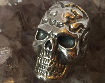 May include: A silver skull ring with intricate details. The skull has a detailed face with a prominent nose and a wide mouth. The ring is set against a background of white crystals.