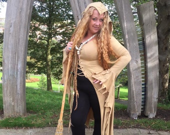 Op de afbeelding: Een vrouw draagt een bruine gekapte tuniek met een lange, vloeiende rok en een zwarte broek. Ze houdt een bezem in haar rechterhand en staat voor een houten constructie.