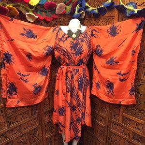 Op de afbeelding: Een oranje jurk met wijde mouwen en een bijpassende riem. De jurk heeft een donkerblauw patroon en wordt op een mannequin gepresenteerd. Een zwarte ketting wordt om de nek van de mannequin gedragen. De jurk is knielang.