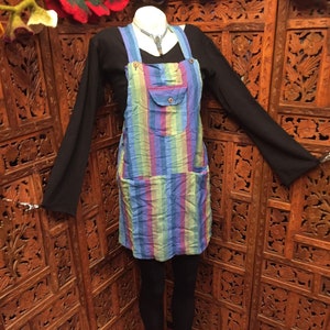 May include: A blue and rainbow striped overall dress with two large pockets. The dress is worn over a black long-sleeved shirt and black leggings. The model is wearing pink polka dot slippers.