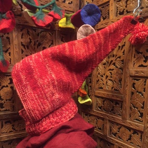 Puede incluir: Una bufanda de punto roja con capucha y un pompón en el extremo. La bufanda está colgada de una pantalla de madera con intrincados tallados.