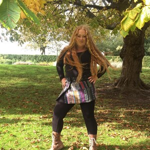 May include: A woman with long, red hair is wearing a black long-sleeved shirt, a colourful patterned skirt, and brown boots. She is standing in a grassy area with fallen leaves.