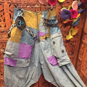 May include: Patchwork harem pants in shades of blue, purple, and olive green. The pants feature a relaxed fit with multiple pockets, decorative stitching, and a black patch with a blue Om symbol. The pants are hanging on a hanger.