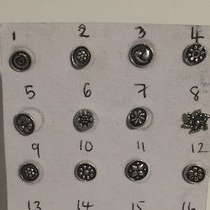 Puede incluir: Una tarjeta blanca con 16 anillos de nariz de plata, cada uno con un diseño diferente, numerados del 1 al 16.