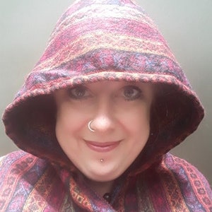 May include: A woman wearing a hooded cloak with a colorful striped pattern. The hood is pulled up over her head, and she is smiling.