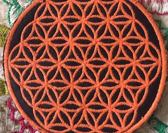 May include: A round, black patch with an orange embroidered flower of life design. The patch is made of felt and has a stitched edge.