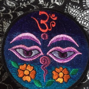 Peut inclure: Patch brodé sur fond bleu, vert et orange. Le patch représente un œil stylisé avec un symbole Om au-dessus. L'œil est entouré de deux fleurs orange.