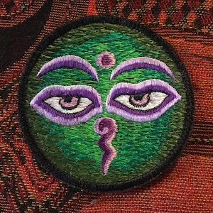 Peut inclure: Patch brodé avec un fond vert et un motif de troisième œil violet. L'œil a un centre blanc et des paupières violettes.