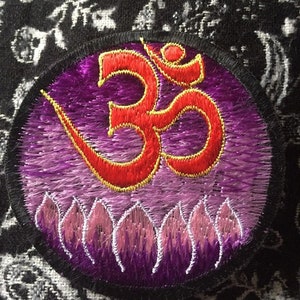 Peut inclure: Un patch brodé circulaire de couleur violet et rouge avec le symbole Om et des fleurs de lotus. Le patch a une bordure noire.
