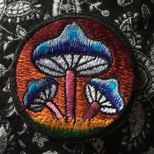 May include: Embroidered patch depicting three stylized mushrooms with blue caps, white gills, and pink stems. The patch has a red and orange background, a black border, and is set against a black paisley fabric.