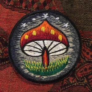 May include: A round, black embroidered patch featuring a white mushroom design. The mushroom has a red cap with yellow dots and a white stem. The mushroom is surrounded by green grass.
