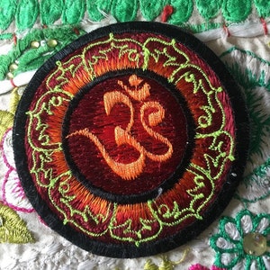 Peut inclure: Un patch rond brodé avec un motif de mandala rouge, orange et vert. Le centre du patch présente un grand symbole Om orange.