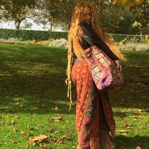 May include: A woman with long, blonde dreadlocks is wearing a long, red, patterned skirt and a black top. She is carrying a large, colourful patchwork bag over her shoulder. The woman is standing in a grassy area with fallen leaves.