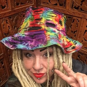 May include: A woman with long blonde dreadlocks is wearing a colourful tie-dye bucket hat with holes. She has multiple facial piercings and is giving a peace sign.