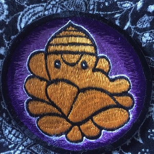 May include: A purple and gold embroidered patch featuring a cartoon octopus with a crown. The octopus is surrounded by a purple circle with a black border.