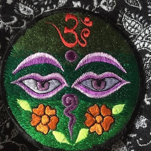 Peut inclure: Patch brodé avec un fond vert et un symbole Om violet. Le patch présente également deux yeux violets avec des pupilles blanches et deux fleurs orange avec des feuilles vertes.