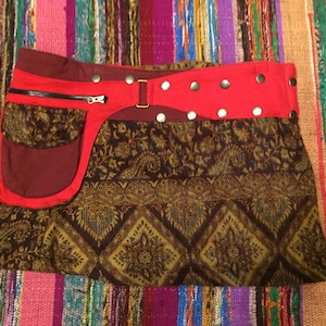 Peut inclure: Une jupe courte à motifs marrons et dorés avec une ceinture rouge et une grande poche rouge sur le côté. La ceinture a une fermeture éclair et des clous argentés.