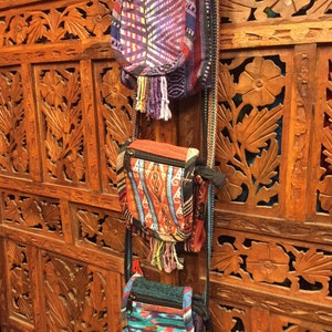 Pode incluir: Tr&ecirc;s pequenas bolsas tecidas coloridas com franjas penduradas em uma parede de madeira com entalhes intrincados. As bolsas t&ecirc;m padr&otilde;es e cores diferentes, incluindo roxo, vermelho, laranja, azul e verde.
