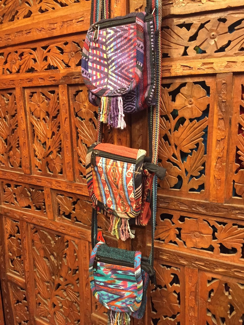 Pode incluir: Tr&ecirc;s pequenas bolsas tecidas coloridas com franjas penduradas em uma parede de madeira com entalhes intrincados. As bolsas t&ecirc;m padr&otilde;es e cores diferentes, incluindo roxo, vermelho, laranja, azul e verde.