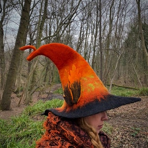 May include: A handmade orange and black witch hat with a pointed crown and wide brim. The hat features a decorative feather design and is made of felt. The hat is worn outdoors in a wooded area.